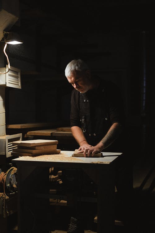 Free Focused male master standing at router table while working with wooden plank in dark carpentry with stack of woods during work process Stock Photo