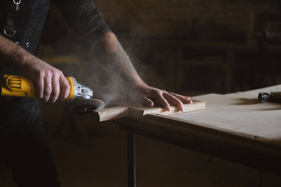 Unrecognizable male master using electric angle grinder to polish lumber plank while standing at table in professional joinery during work