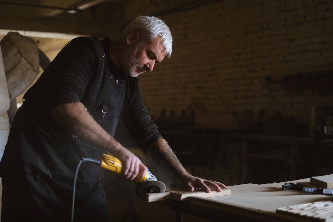 Side view of skilled male master in apron using electric angle grinder to polish wooden plank at table in professional carpentry