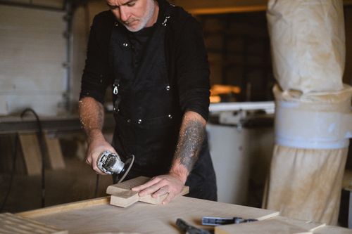 Free Focused male master in apron with angle grinder working with wooden board while standing at table with special instruments in workshop Stock Photo