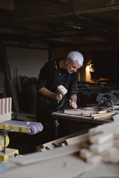Side view of skilled male carpenter using mallet and chisel while creating pattern on wooden detail during work in joinery with blurred boards