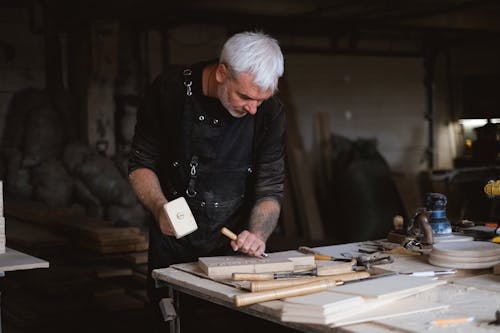 Serious male woodworker in apron using mallet and chisel to carve wooden board at table with abundance of instruments in carpentry