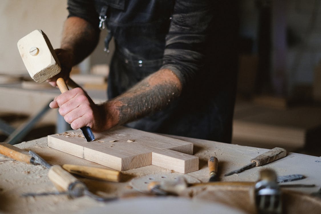 Unrecognizable male carpenter using forged chisel and wooden hammer while creating pattern on lumber board at table in workshop on blurred background