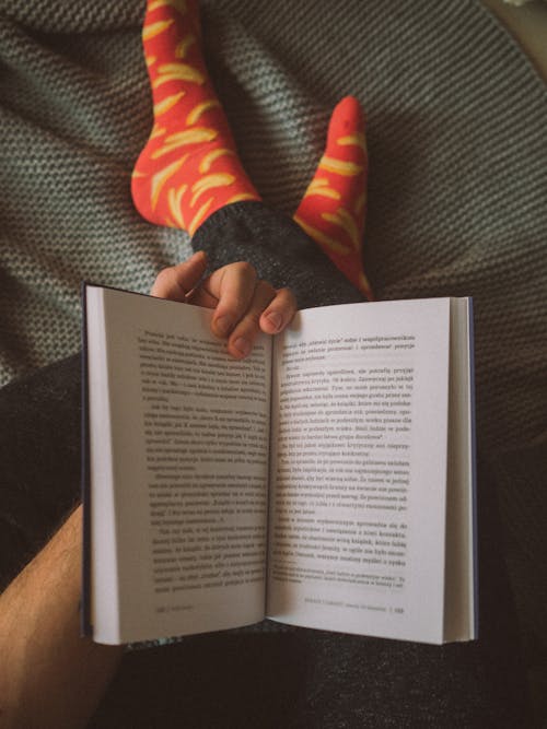 Free From above of crop unrecognizable male in casual clothes and colorful socks sitting on comfy bed and reading interesting book during weekend at home Stock Photo
