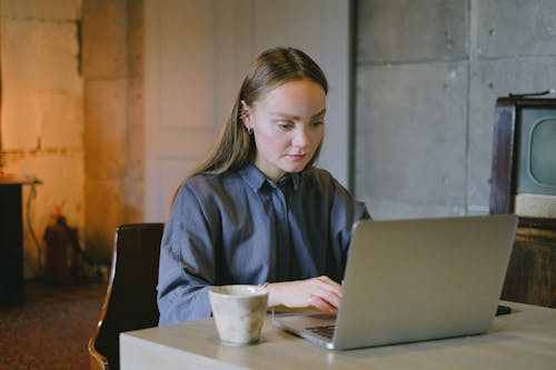 Concentrated young lady in casual outfit sitting on chair at table and working remotely on laptop near cup of drink in loft