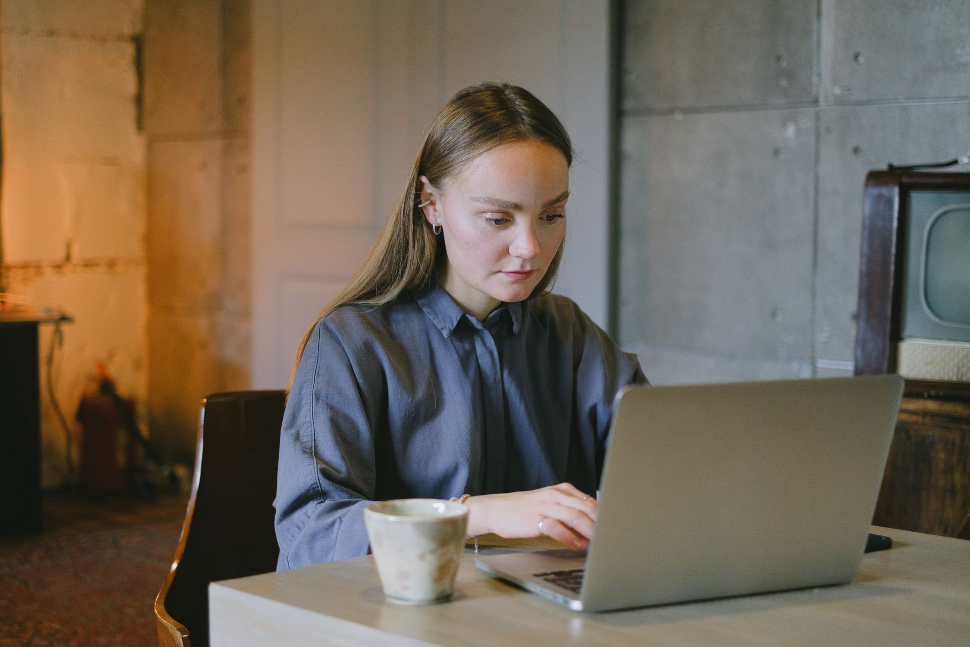 Concentrated young lady in casual outfit sitting on chair at table and working remotely on laptop near cup of drink in loft