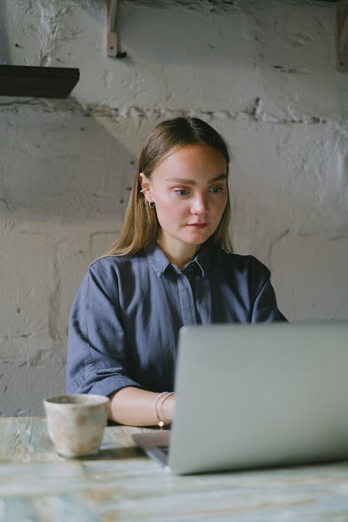 Free Serious young woman in casual clothes sitting at table and working remotely on netbook near mug with drink in workplace Stock Photo