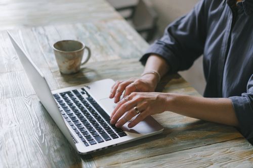 Free Crop faceless female freelancer in casual outfit sitting at table with cup of drink and surfing on laptop in light workspace Stock Photo