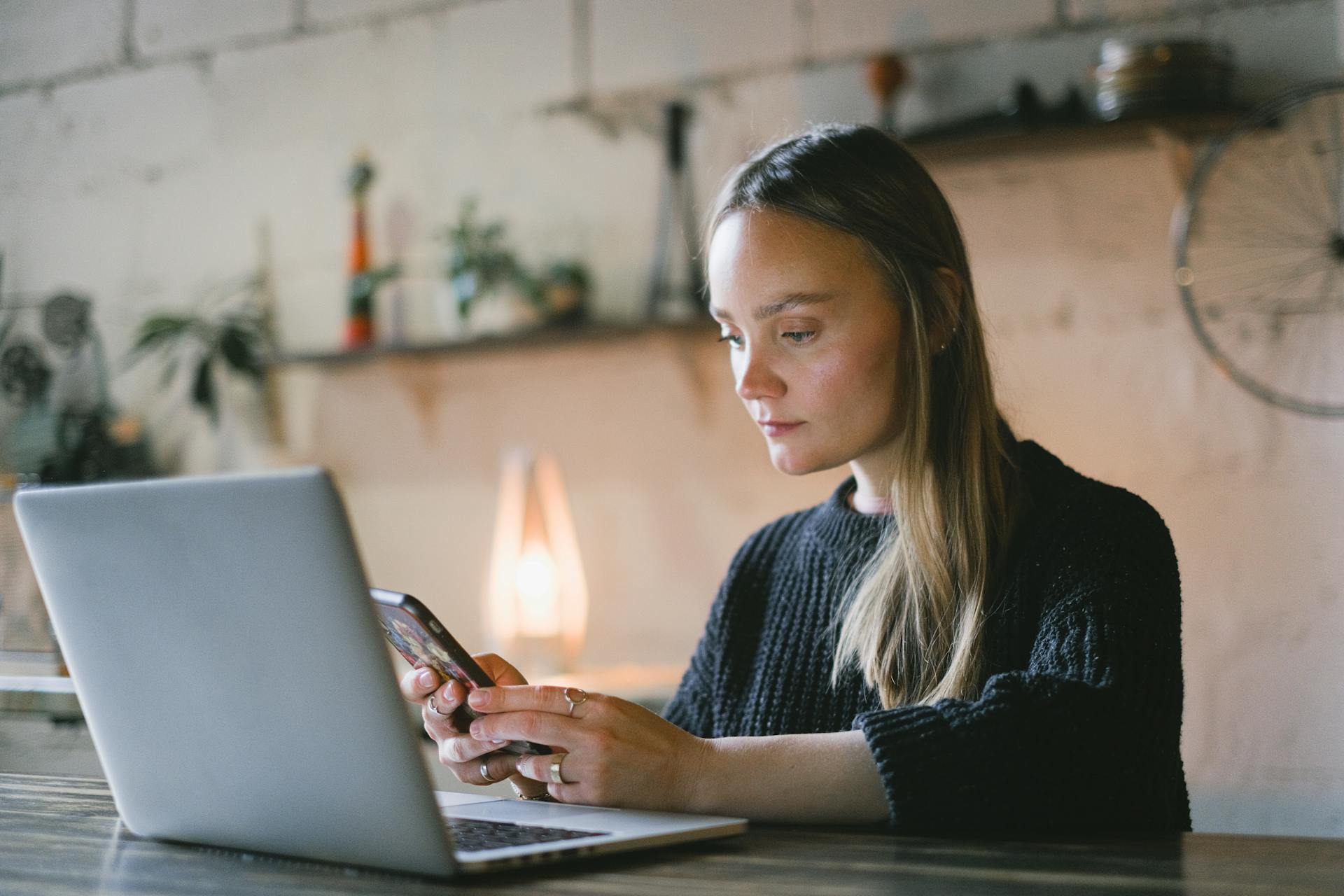 Concentrated female freelancer text messaging on cellphone while sitting at table with netbook during online work on blurred background