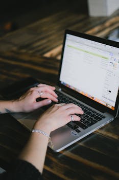 A woman is typing on a laptop at a wooden desk, focused on work in a modern indoor setting.