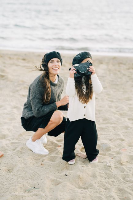 A mother and daughter bond on the beach as the young girl playfully uses a camera.