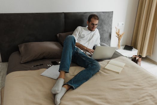 Adult man working on a laptop while sitting on bed at home, casual and relaxed environment.