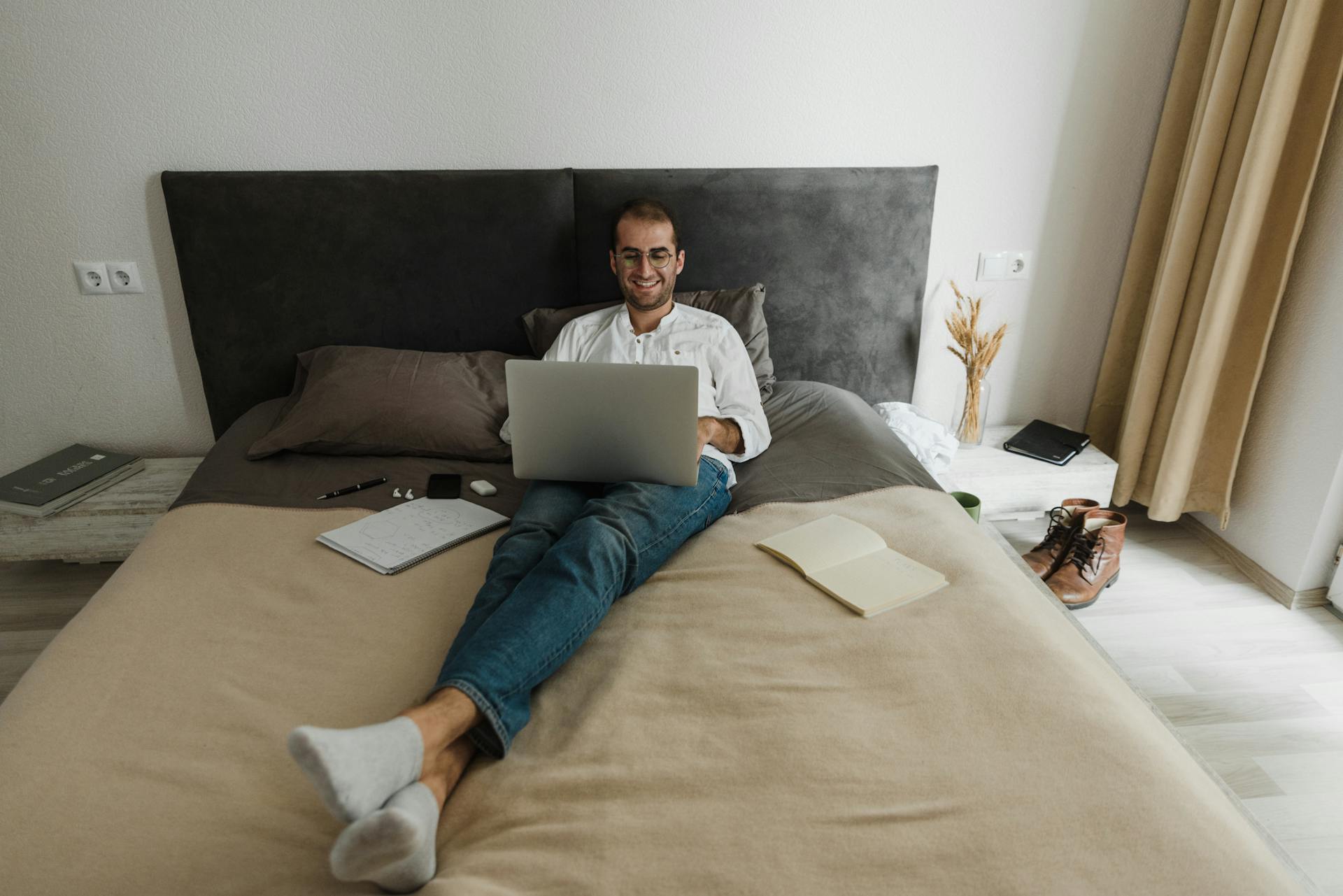 A smiling man working remotely on his laptop in a cozy bedroom setting, embracing a modern lifestyle.