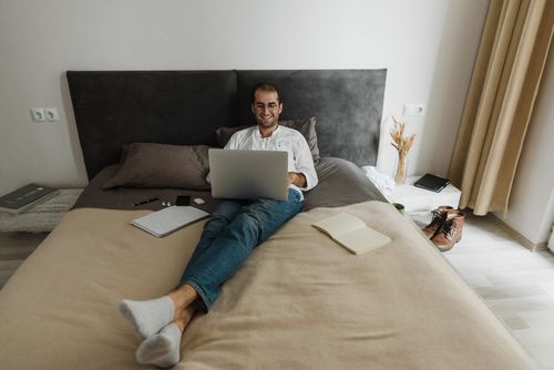 Free A smiling man working remotely on his laptop in a cozy bedroom setting, embracing a modern lifestyle. Stock Photo
