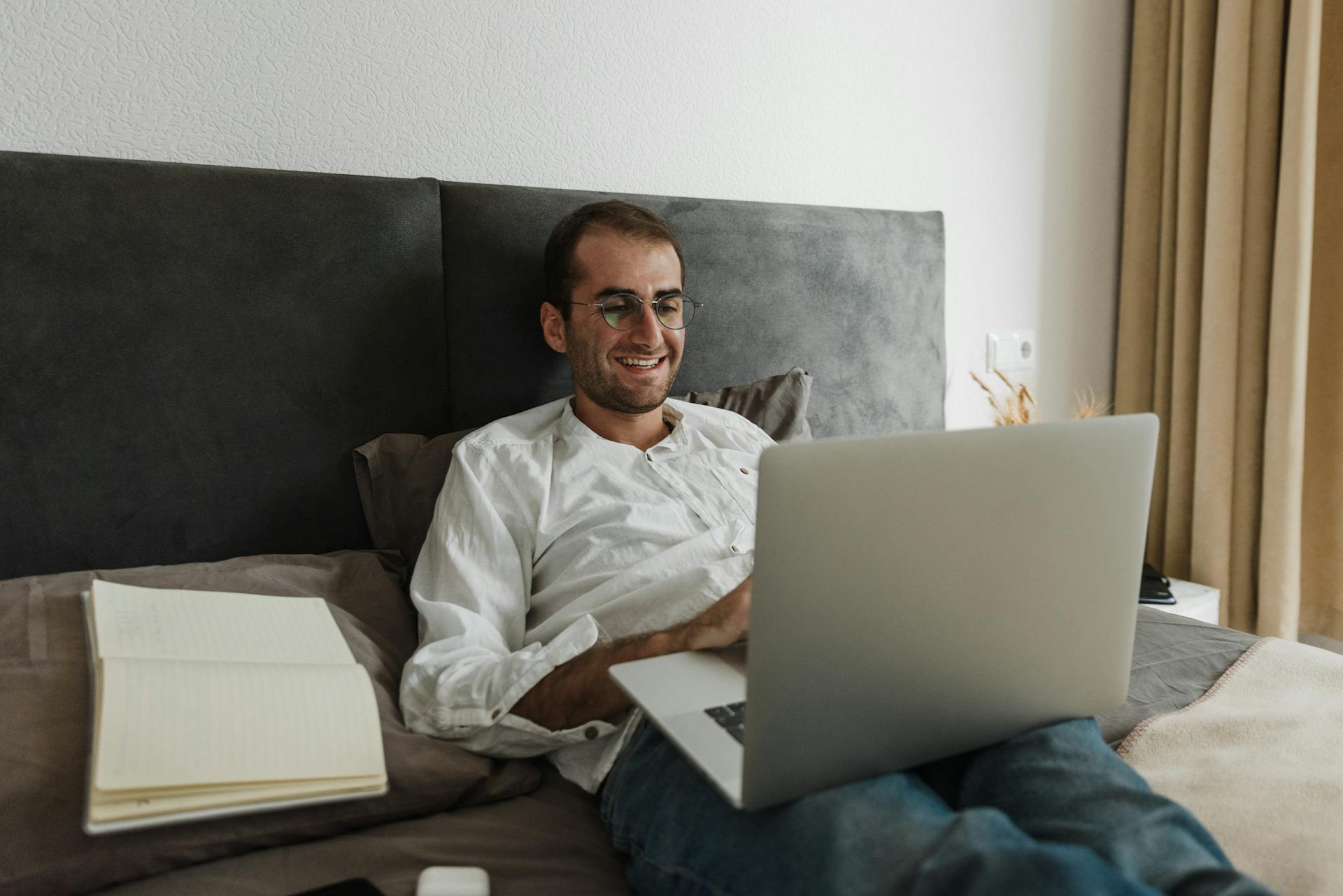 Smiling man in glasses working on laptop from bed with a notebook beside him.