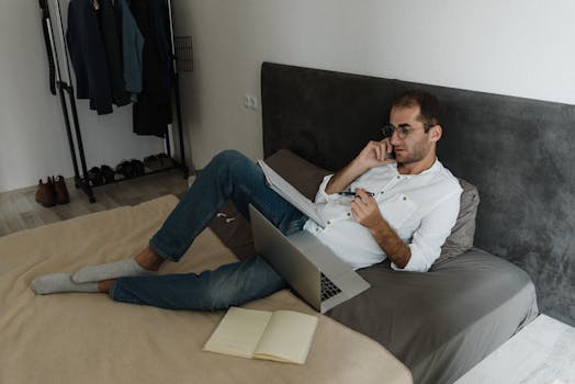 A man multitasking with a laptop and phone on a bed, creating a relaxed home office setup.