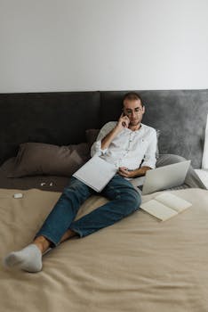 Man relaxing on bed, using laptop and phone, working from home setup.