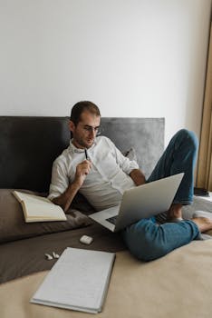 Man in white shirt working remotely on laptop from bed, demonstrating modern work-life balance.