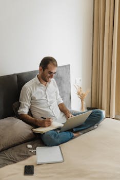 Man working from home on a laptop, seated on bed while writing notes and smiling.