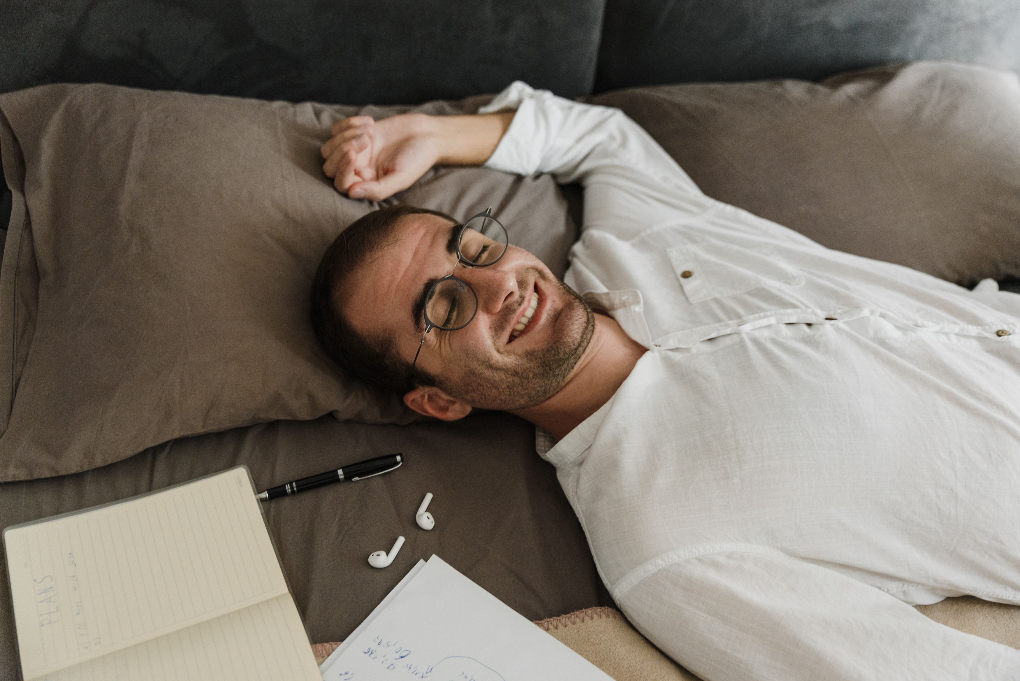 Man in white shirt smiling and relaxing in bed with notes and headphones nearby.