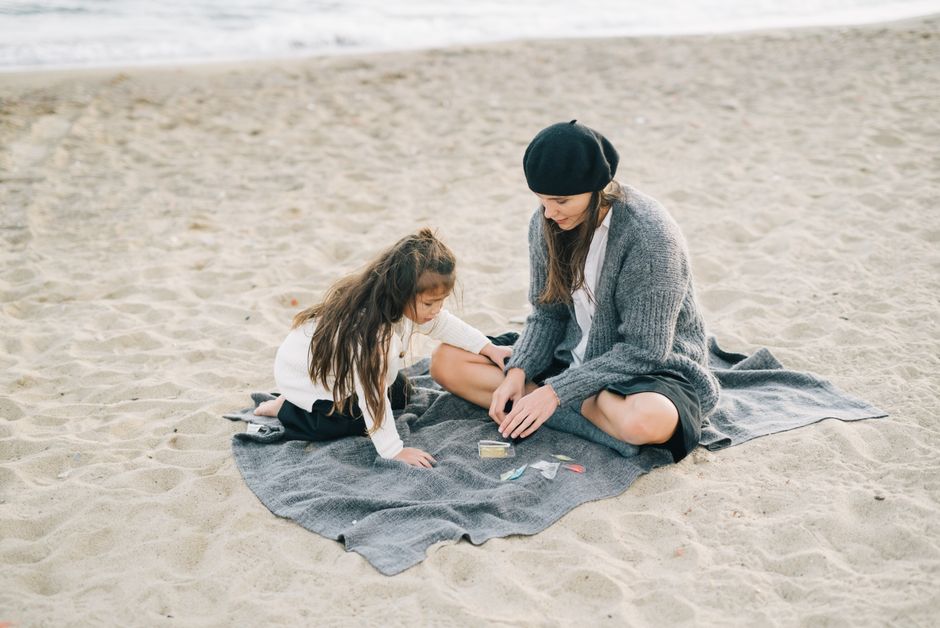 A mother and daughter enjoy quality time together on a sandy beach, sitting on a blanket.