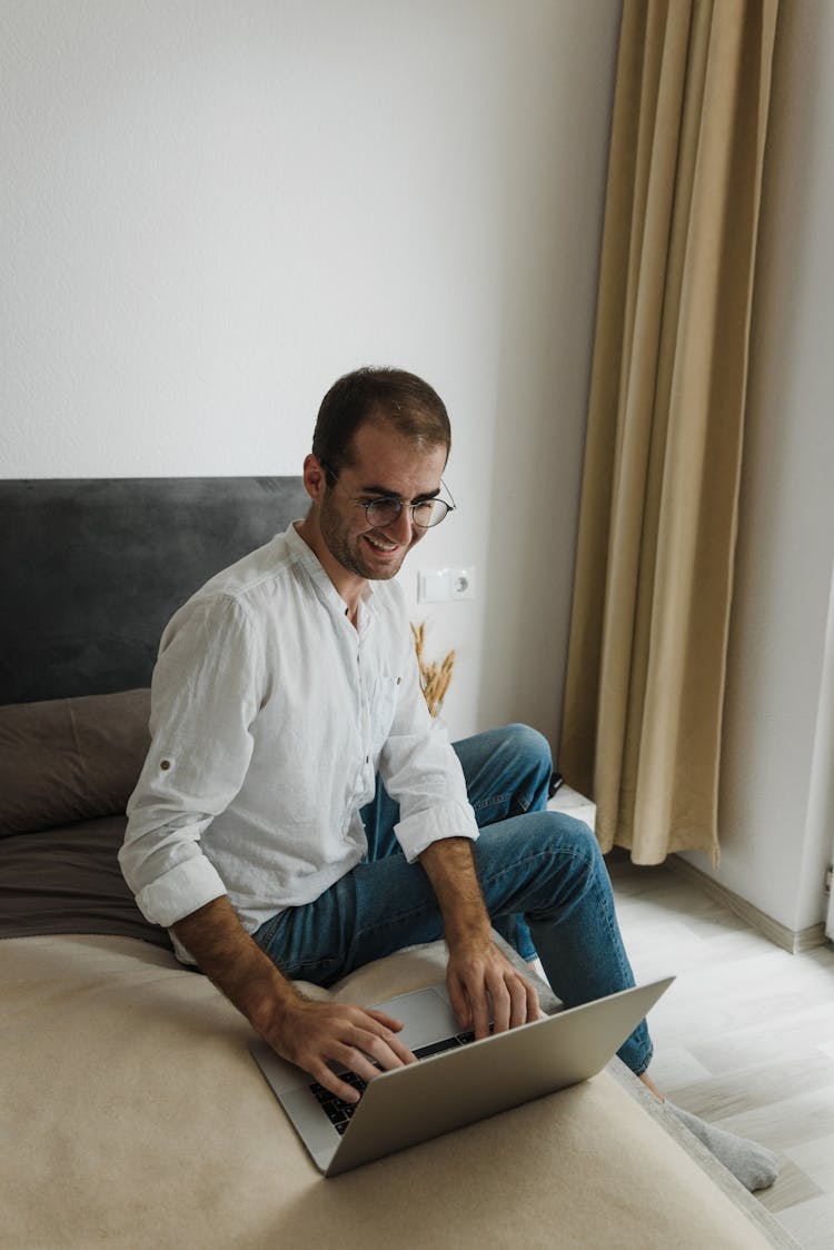 A Man In White Long Sleeves Sitting On The Side Of The Bed While Typing On A Laptop