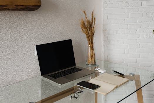 A sleek home office setup featuring a laptop, smartphone, and notepad on a glass desk.