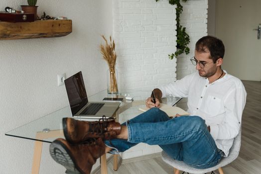 Man in relaxed pose writing in notebook with feet up in home office setting.