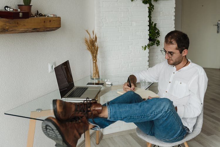 A Man Writing On A Notebook With Feet On Top Of Desk