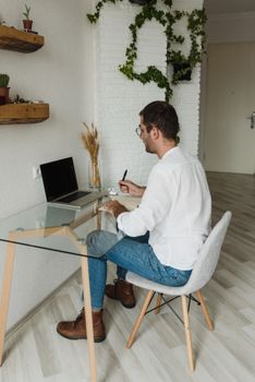 A man sits at a glass desk working on a laptop in a modern, plant-decorated home office.