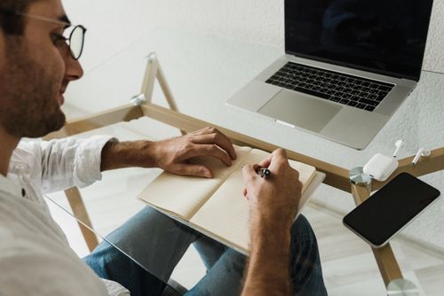 Free Person writing in a notebook while sitting at a modern glass desk with a laptop and smartphone. Stock Photo