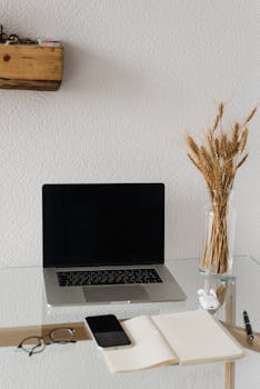 A modern minimalist workspace featuring a laptop, notebook, and a vase of dried wheat on a glass table.