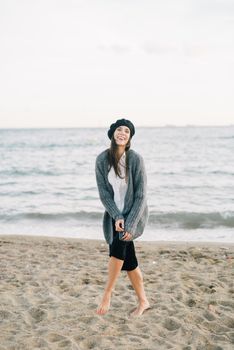 一名赤脚站在沙滩上的女人，穿着羊毛衫、戴着帽子，笑着