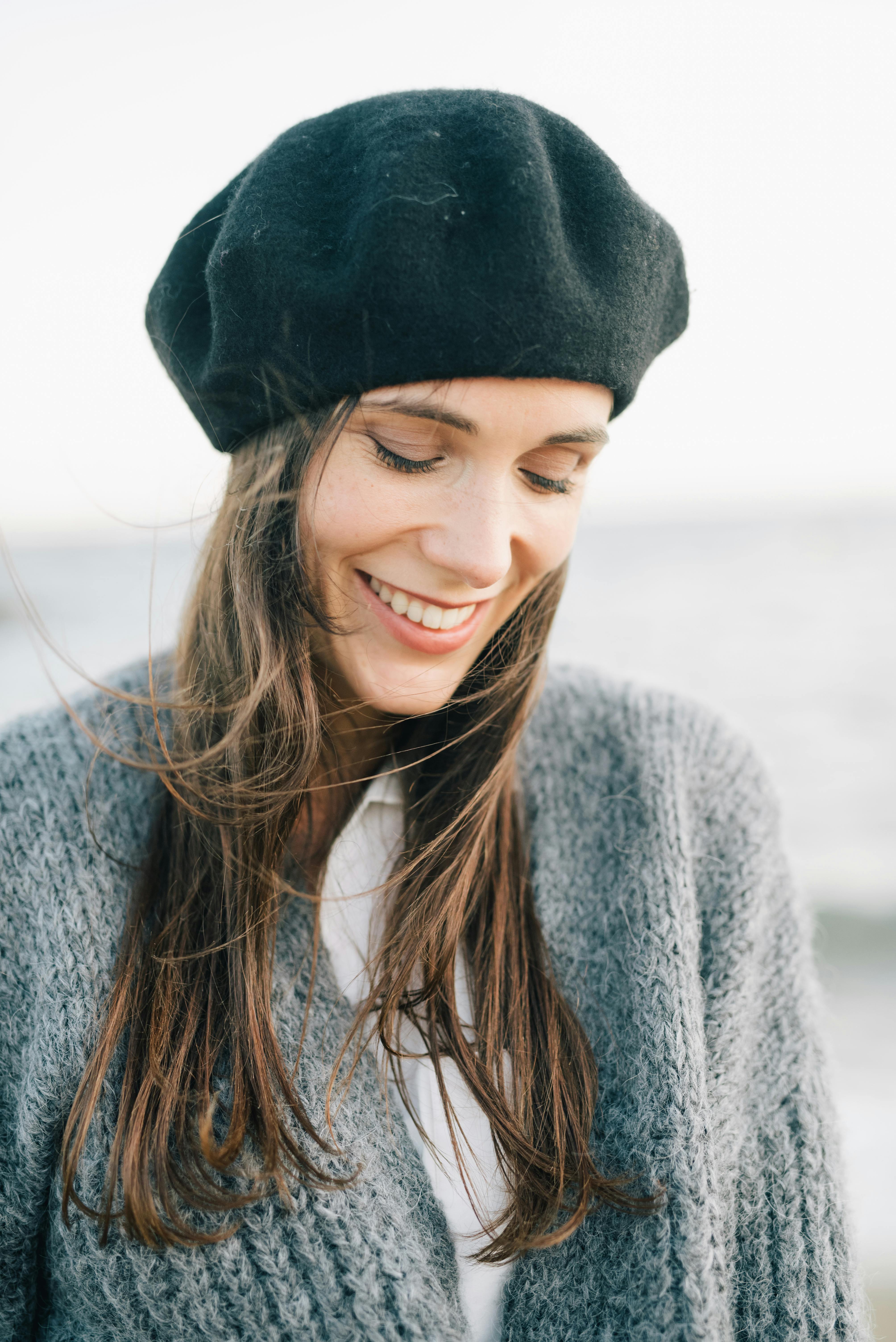 Portrait of a woman smiling with a gentle breeze at the beach, wearing a black hat and gray sweater.