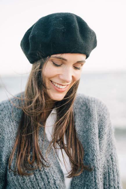 Portrait of a woman smiling with a gentle breeze at the beach, wearing a black hat and gray sweater.