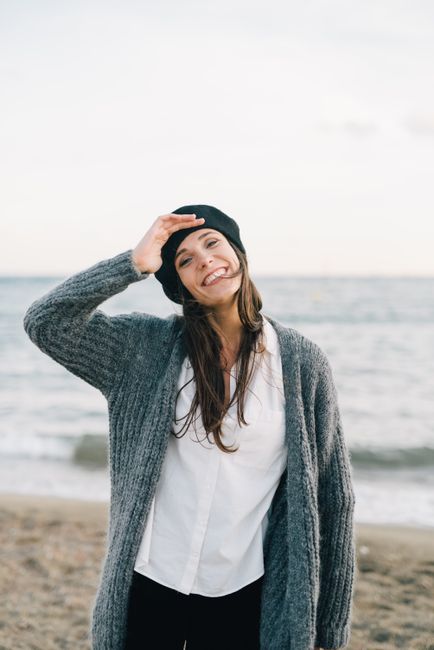 A joyful woman in a knitted cardigan stands on the beach, smiling warmly.