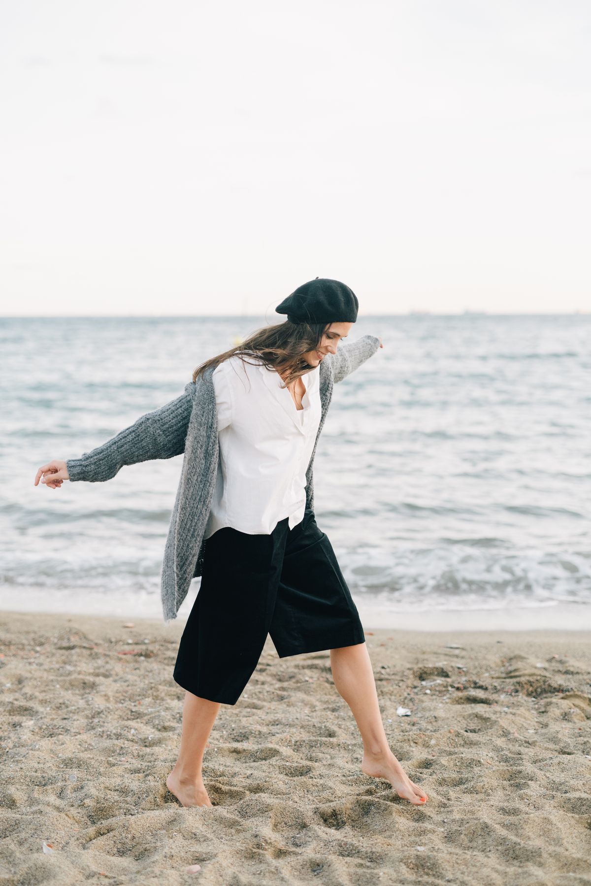 A woman in a white shirt and grey knit layered on the beach — the fine knit, layered