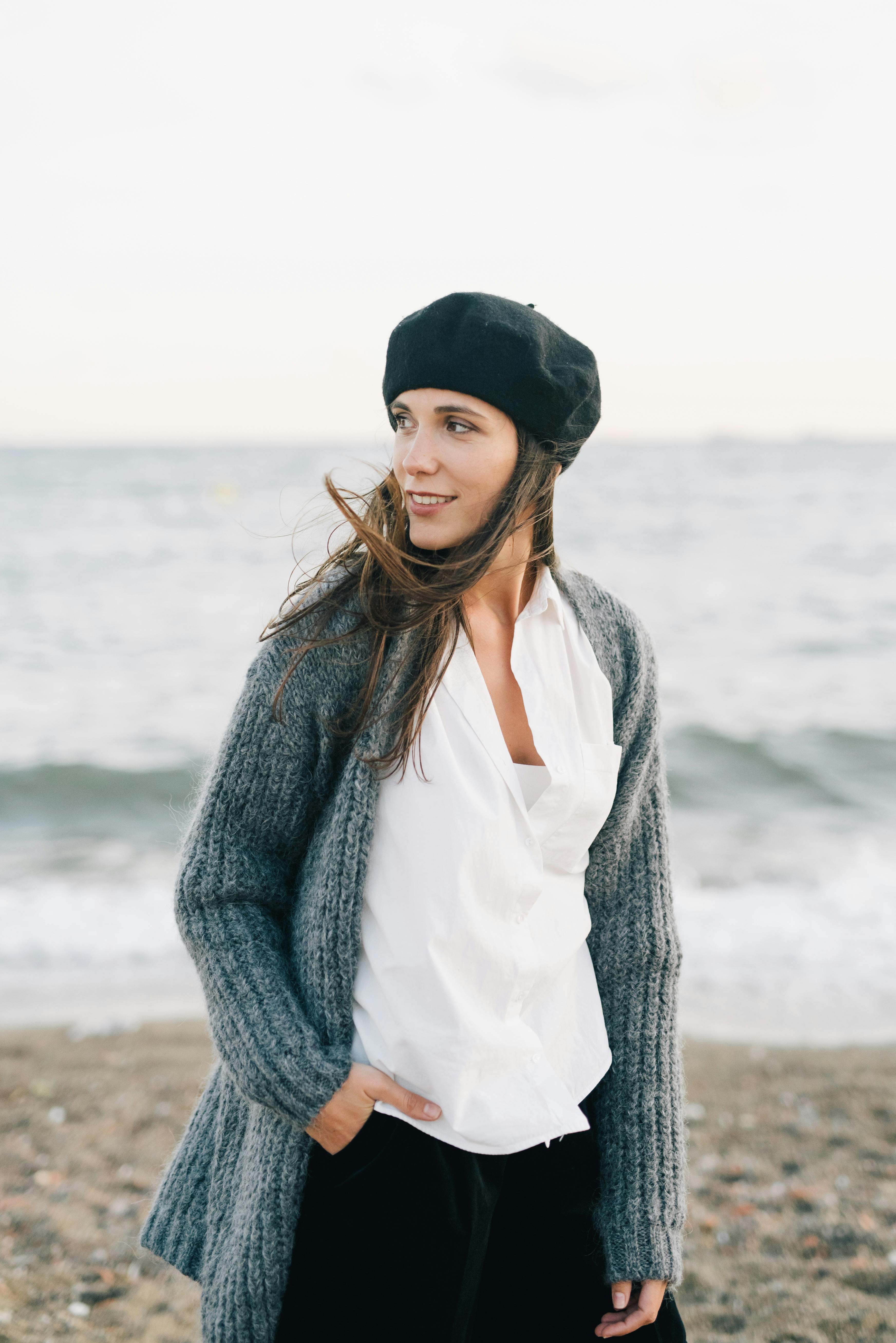 Young woman in a gray sweater and beret enjoying a breezy beach day.