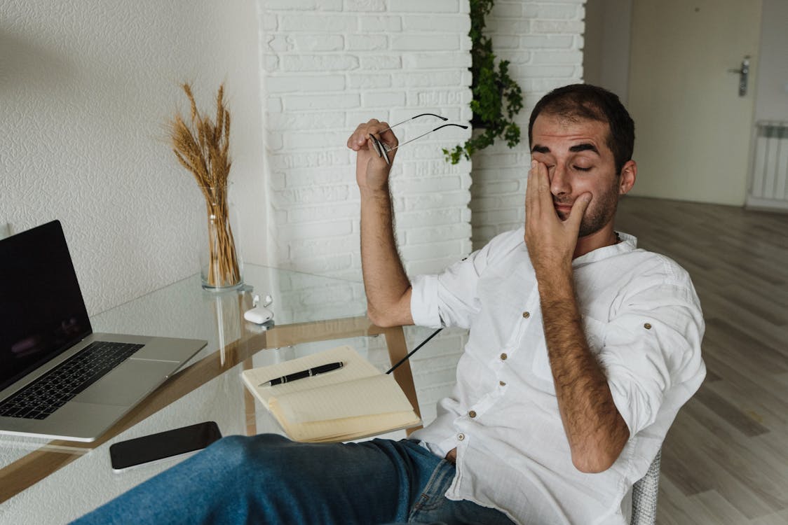 Free A tired man in a white shirt rests at a home office desk, holding eyeglasses and rubbing his eyes. Stock Photo