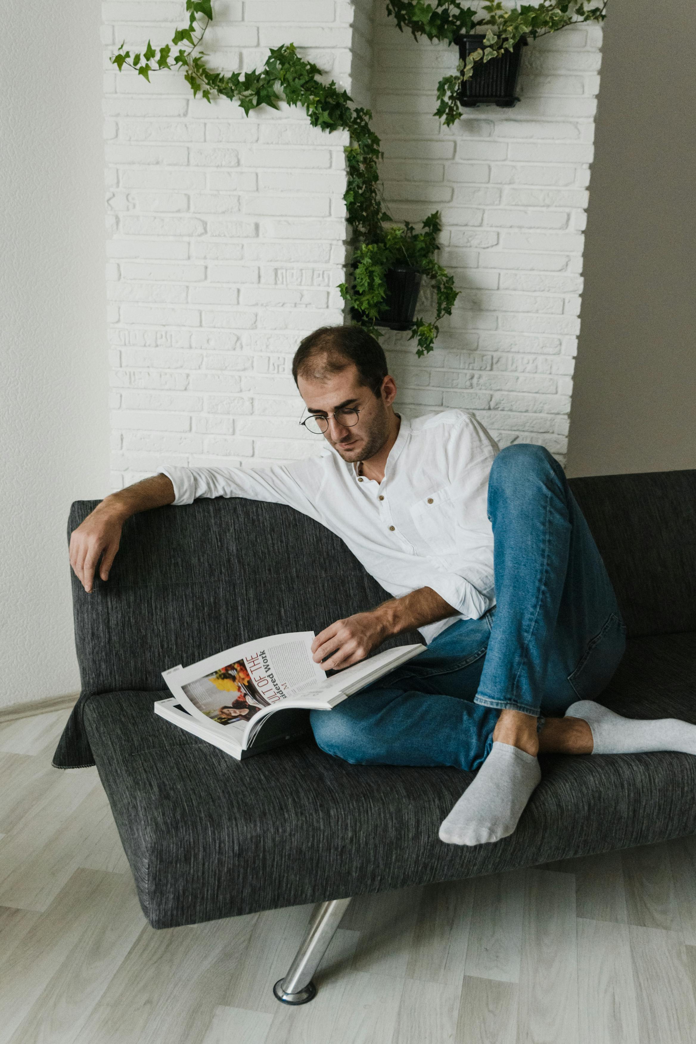man in white polo shirt sitting on gray couch reading book