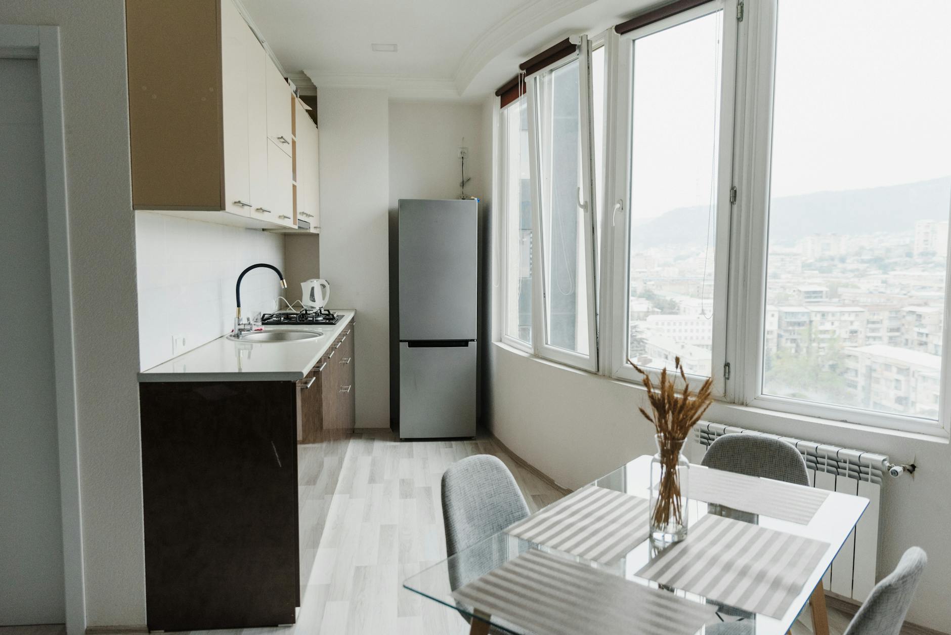 Bright modern kitchen with a glass table, minimalist design, and cityscape view through large windows.