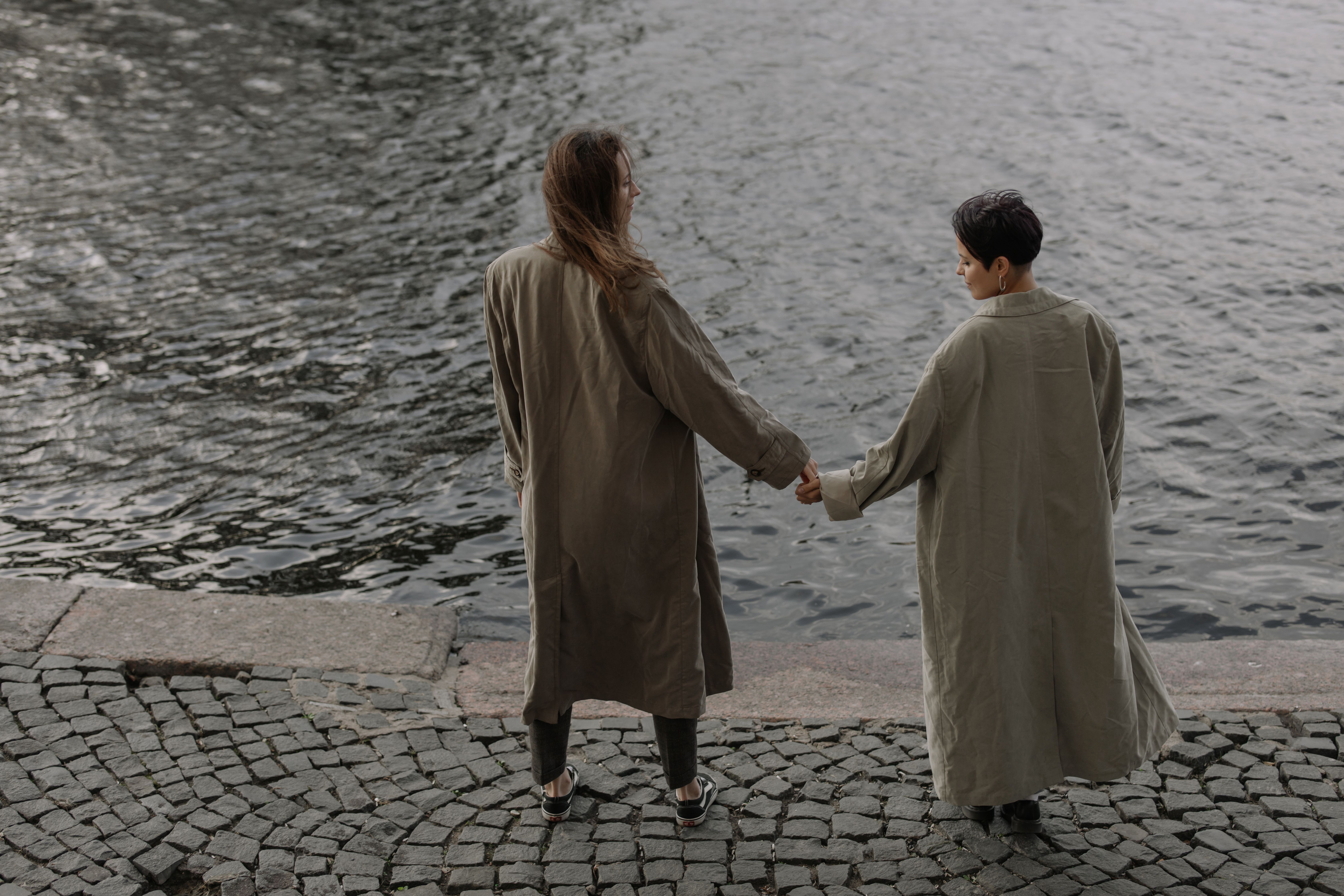Two women holding hands by a riverside, wearing trench coats, symbolizing love and togetherness.