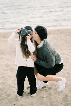 A mother kisses her daughter taking photos on a sandy beach.