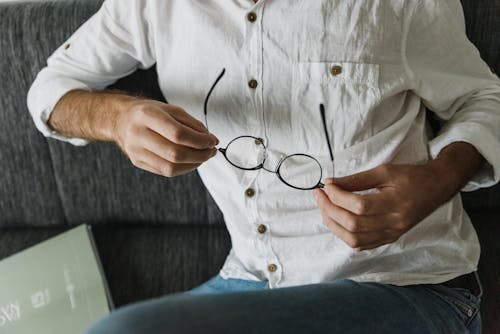 From above of crop unrecognizable male wearing casual stylish outfit with shirt and denim pants sitting with glasses in hands