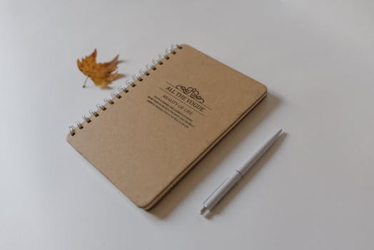 A simple autumn-themed flat lay of a notebook with a pen and dried leaf on a white background.