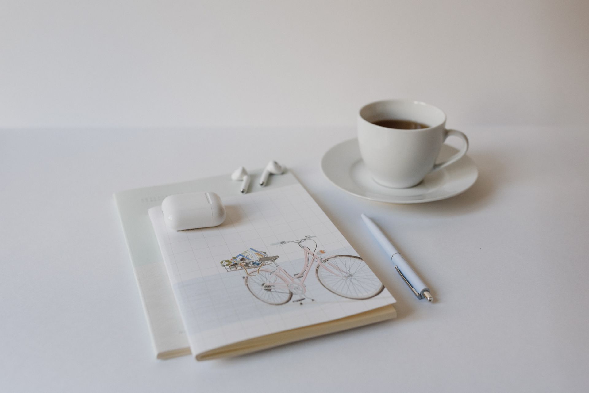 A minimalist setup with a cup of coffee, notepad, pen, and wireless earbuds on a white background.