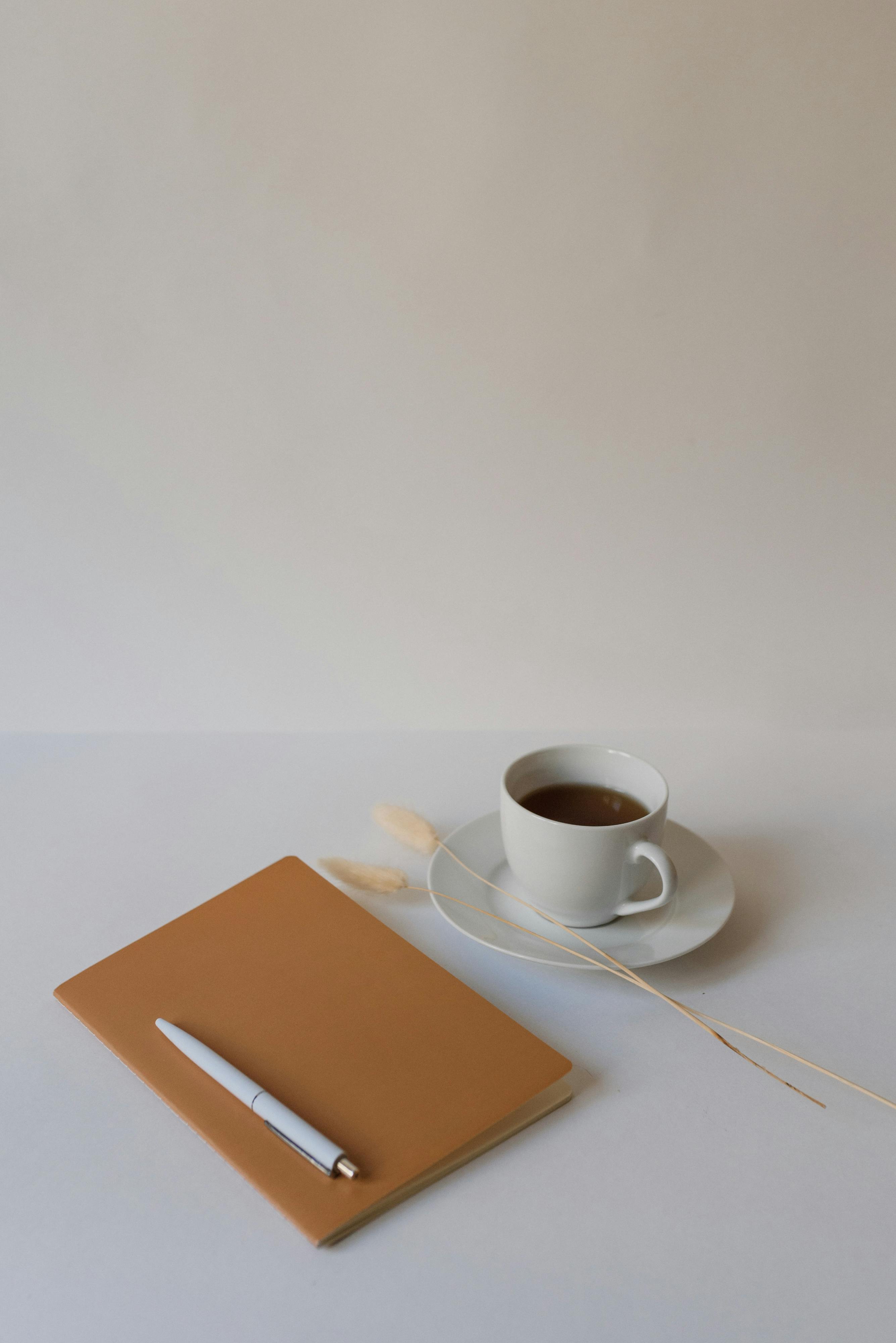 白色表面上呈现着咖啡杯、记事本和笔的极简主义场景