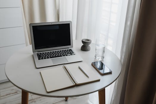 Compact minimalist workspace featuring a laptop, smartphone, notebook, glass and candle holder.