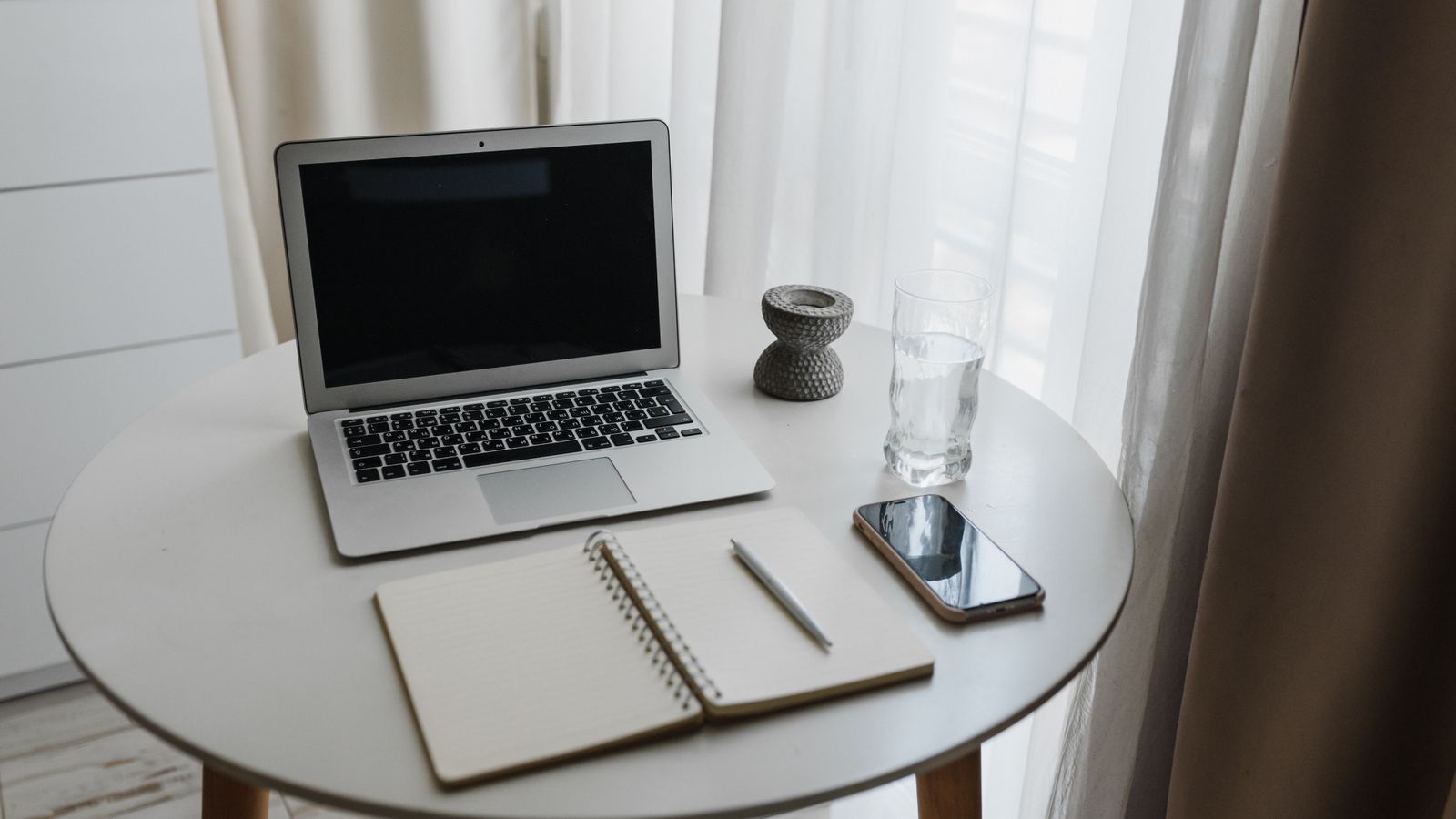 A notebook, phone and laptop on a table beside a window
