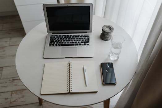 A neat workspace featuring a laptop, notebook, pen, and smartphone on a round table indoors.